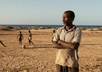 Entrenador de Cabo Verde en campo de tierra bajo el sol, con niños jugando al fútbol con un balón improvisado