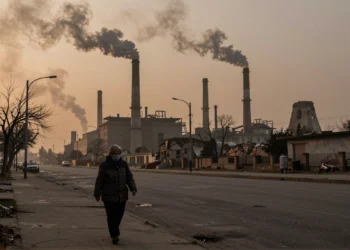 De las hogueras prehistóricas al aire irrespirable: cómo la contaminación se convirtió en la mayor amenaza ambiental para la salud humana 1 Anciana con mascarilla camina bajo cielos contaminados frente a chimeneas industriales humeantes al atardecer